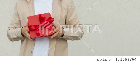 Man giving red gift box tied with ribbon against beige wall. Birthday present. Valentine's Day. Banner. Space for design. 131068864