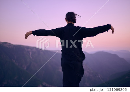 Woman practicing Tai Chi in traditional black outfit on mountain at sunrise, arms outstretched in meditation pose focusing on balance, energy, and mindfulness in nature. 131069206