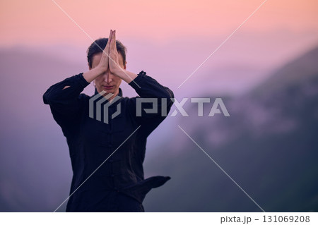 Woman practicing Tai Chi in traditional black outfit on mountain at sunrise, arms outstretched in meditation pose focusing on balance, energy, and mindfulness in nature. 131069208