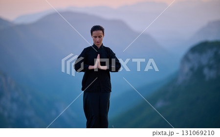 Woman practicing Tai Chi in traditional black outfit on mountain at sunrise, arms outstretched in meditation pose focusing on balance, energy, and mindfulness in nature. 131069210