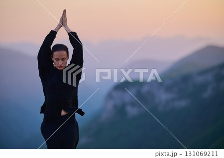 Woman practicing Tai Chi in traditional black outfit on mountain at sunrise, arms outstretched in meditation pose focusing on balance, energy, and mindfulness in nature. 131069211
