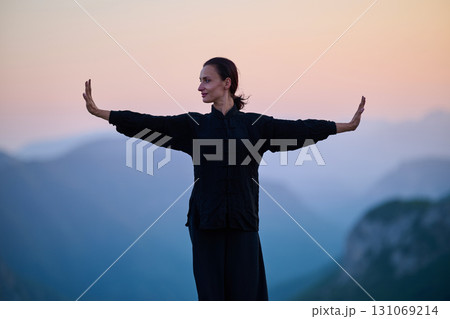 Woman practicing Tai Chi in traditional black outfit on mountain at sunrise, arms outstretched in meditation pose focusing on balance, energy, and mindfulness in nature. 131069214