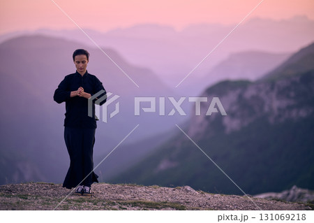 Woman practicing Tai Chi in traditional black outfit on mountain at sunrise, arms outstretched in meditation pose focusing on balance, energy, and mindfulness in nature. 131069218
