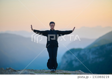 Woman practicing Tai Chi in traditional black outfit on mountain at sunrise, arms outstretched in meditation pose focusing on balance, energy, and mindfulness in nature. Woman practicing Tai Chi in traditional black outfit on mountain at sunrise, arms outstretched in meditation pose focusing on balance, energy, and mindfulness in nature. 131069219
