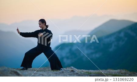 Woman practicing Tai Chi in traditional black outfit on mountain at sunrise, arms outstretched in meditation pose focusing on balance, energy, and mindfulness in nature. 131069220