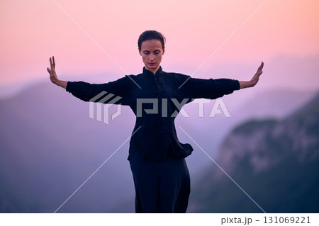 Woman practicing Tai Chi in traditional black outfit on mountain at sunrise, arms outstretched in meditation pose focusing on balance, energy, and mindfulness in nature. Woman practicing Tai Chi in traditional black outfit on mountain at sunrise, arms outstretched in meditation pose focusing on balance, energy, and mindfulness in nature. 131069221