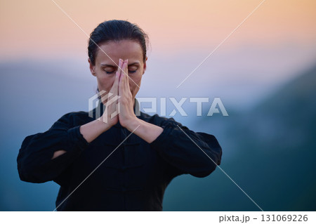 Woman practicing Tai Chi in traditional black outfit on mountain at sunrise, arms outstretched in meditation pose focusing on balance, energy, and mindfulness in nature. 131069226