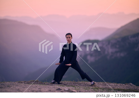 Woman practicing Tai Chi in traditional black outfit on mountain at sunrise, arms outstretched in meditation pose focusing on balance, energy, and mindfulness in nature. 131069246