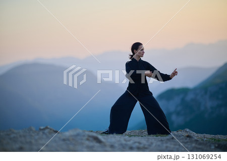 Woman practicing Tai Chi in traditional black outfit on mountain at sunrise, arms outstretched in meditation pose focusing on balance, energy, and mindfulness in nature. 131069254