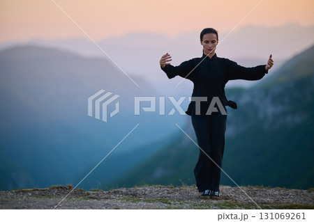 Woman practicing Tai Chi in traditional black outfit on mountain at sunrise, arms outstretched in meditation pose focusing on balance, energy, and mindfulness in nature. 131069261