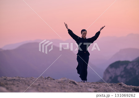 Woman practicing Tai Chi in traditional black outfit on mountain at sunrise, arms outstretched in meditation pose focusing on balance, energy, and mindfulness in nature. 131069262