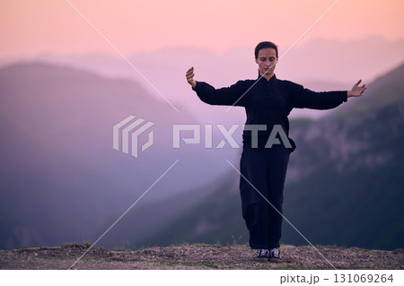 Woman practicing Tai Chi in traditional black outfit on mountain at sunrise, arms outstretched in meditation pose focusing on balance, energy, and mindfulness in nature. 131069264