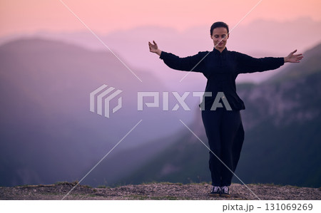Woman practicing Tai Chi in traditional black outfit on mountain at sunrise, arms outstretched in meditation pose focusing on balance, energy, and mindfulness in nature. 131069269