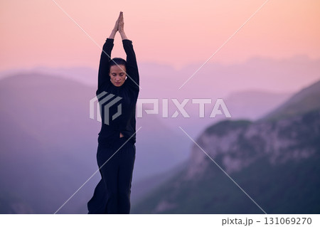 Woman practicing Tai Chi in traditional black outfit on mountain at sunrise, arms outstretched in meditation pose focusing on balance, energy, and mindfulness in nature. 131069270