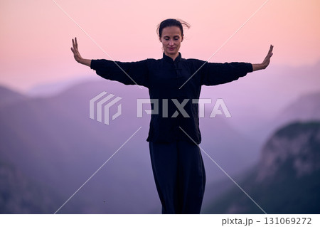 Woman practicing Tai Chi in traditional black outfit on mountain at sunrise, arms outstretched in meditation pose focusing on balance, energy, and mindfulness in nature. 131069272