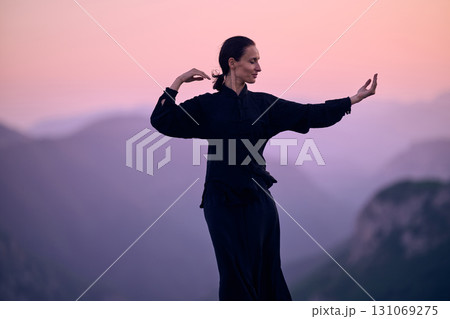 Woman practicing Tai Chi in traditional black outfit on mountain at sunrise, arms outstretched in meditation pose focusing on balance, energy, and mindfulness in nature. 131069275
