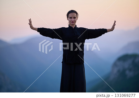 Woman practicing Tai Chi in traditional black outfit on mountain at sunrise, arms outstretched in meditation pose focusing on balance, energy, and mindfulness in nature. 131069277