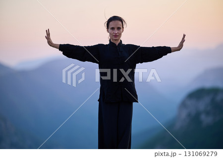 Woman practicing Tai Chi in traditional black outfit on mountain at sunrise, arms outstretched in meditation pose focusing on balance, energy, and mindfulness in nature. 131069279