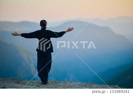 Woman practicing Tai Chi in traditional black outfit on mountain at sunrise, arms outstretched in meditation pose focusing on balance, energy, and mindfulness in nature. 131069281