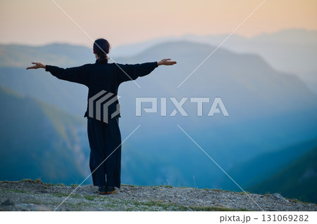 Woman practicing Tai Chi in traditional black outfit on mountain at sunrise, arms outstretched in meditation pose focusing on balance, energy, and mindfulness in nature. 131069282