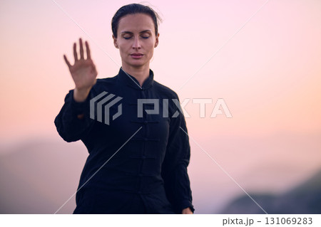 Woman practicing Tai Chi in traditional black outfit on mountain at sunrise, arms outstretched in meditation pose focusing on balance, energy, and mindfulness in nature. 131069283