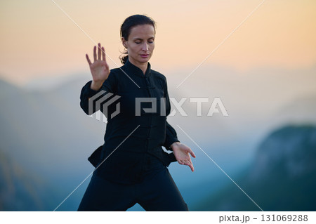 Woman practicing Tai Chi in traditional black outfit on mountain at sunrise, arms outstretched in meditation pose focusing on balance, energy, and mindfulness in nature. Woman practicing Tai Chi in traditional black outfit on mountain at sunrise, arms outstretched in meditation pose focusing on balance, energy, and mindfulness in nature. 131069288