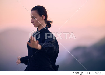 Woman practicing Tai Chi in traditional black outfit on mountain at sunrise, arms outstretched in meditation pose focusing on balance, energy, and mindfulness in nature. 131069290
