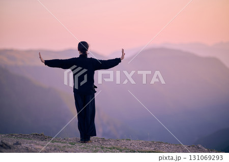 Woman practicing Tai Chi in traditional black outfit on mountain at sunrise, arms outstretched in meditation pose focusing on balance, energy, and mindfulness in nature. Woman practicing Tai Chi in traditional black outfit on mountain at sunrise, arms outstretched in meditation pose focusing on balance, energy, and mindfulness in nature. 131069293