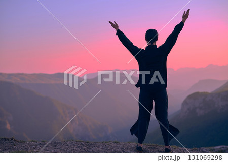 Woman practicing Tai Chi in traditional black outfit on mountain at sunrise, arms outstretched in meditation pose focusing on balance, energy, and mindfulness in nature. 131069298