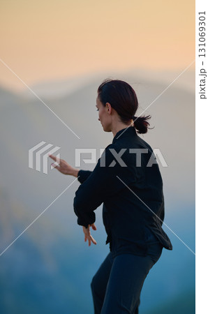 Woman practicing Tai Chi in traditional black outfit on mountain at sunrise, arms outstretched in meditation pose focusing on balance, energy, and mindfulness in nature. 131069301