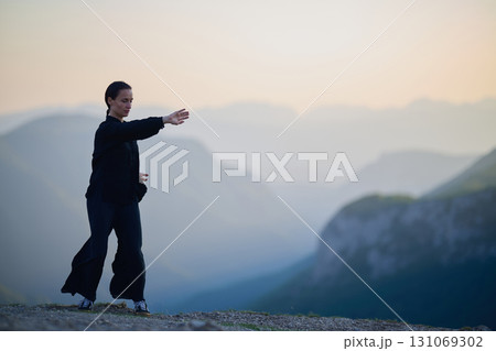 Woman practicing Tai Chi in traditional black outfit on mountain at sunrise, arms outstretched in meditation pose focusing on balance, energy, and mindfulness in nature. 131069302