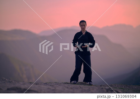 Woman practicing Tai Chi in traditional black outfit on mountain at sunrise, arms outstretched in meditation pose focusing on balance, energy, and mindfulness in nature. 131069304