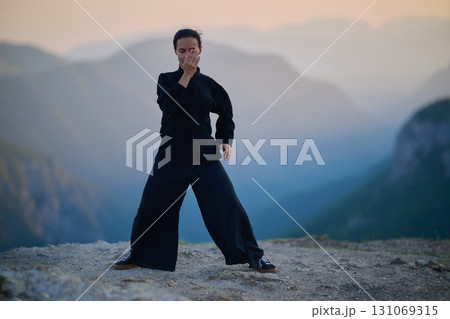 Woman practicing Tai Chi in traditional black outfit on mountain at sunrise, arms outstretched in meditation pose focusing on balance, energy, and mindfulness in nature. Woman practicing Tai Chi in traditional black outfit on mountain at sunrise, arms outstretched in meditation pose focusing on balance, energy, and mindfulness in nature. 131069315