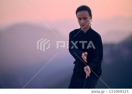 Woman practicing Tai Chi in traditional black outfit on mountain at sunrise, arms outstretched in meditation pose focusing on balance, energy, and mindfulness in nature. 131069325