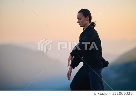 Woman practicing Tai Chi in traditional black outfit on mountain at sunrise, arms outstretched in meditation pose focusing on balance, energy, and mindfulness in nature. Woman practicing Tai Chi in traditional black outfit on mountain at sunrise, arms outstretched in meditation pose focusing on balance, energy, and mindfulness in nature. 131069329