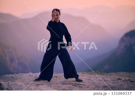 Woman practicing Tai Chi in traditional black outfit on mountain at sunrise, arms outstretched in meditation pose focusing on balance, energy, and mindfulness in nature. Woman practicing Tai Chi in traditional black outfit on mountain at sunrise, arms outstretched in meditation pose focusing on balance, energy, and mindfulness in nature. 131069337