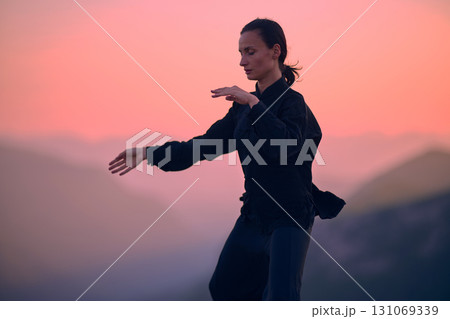 Woman practicing Tai Chi in traditional black outfit on mountain at sunrise, arms outstretched in meditation pose focusing on balance, energy, and mindfulness in nature. 131069339