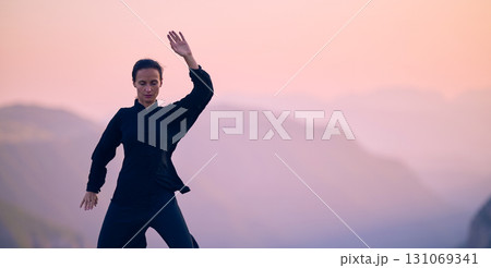 Woman practicing Tai Chi in traditional black outfit on mountain at sunrise, arms outstretched in meditation pose focusing on balance, energy, and mindfulness in nature. 131069341