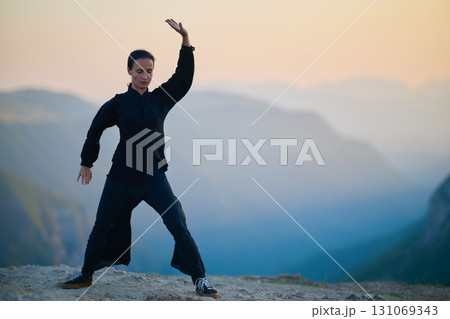 Woman practicing Tai Chi in traditional black outfit on mountain at sunrise, arms outstretched in meditation pose focusing on balance, energy, and mindfulness in nature. 131069343