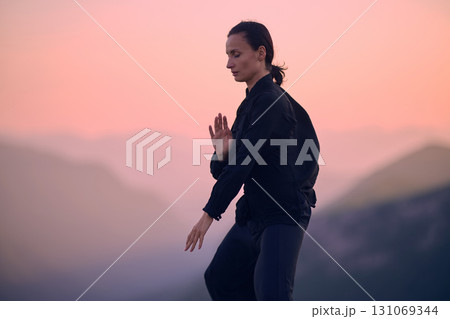Woman practicing Tai Chi in traditional black outfit on mountain at sunrise, arms outstretched in meditation pose focusing on balance, energy, and mindfulness in nature. Woman practicing Tai Chi in traditional black outfit on mountain at sunrise, arms outstretched in meditation pose focusing on balance, energy, and mindfulness in nature. 131069344