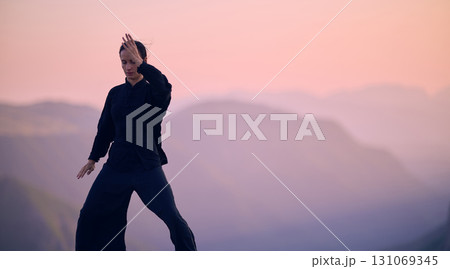 Woman practicing Tai Chi in traditional black outfit on mountain at sunrise, arms outstretched in meditation pose focusing on balance, energy, and mindfulness in nature. 131069345