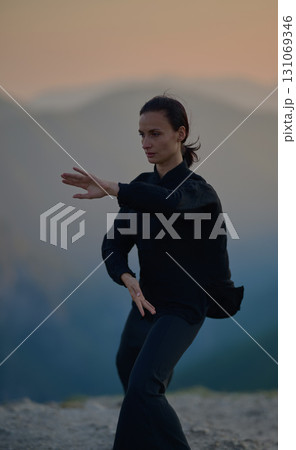 Woman practicing Tai Chi in traditional black outfit on mountain at sunrise, arms outstretched in meditation pose focusing on balance, energy, and mindfulness in nature. Woman practicing Tai Chi in traditional black outfit on mountain at sunrise, arms outstretched in meditation pose focusing on balance, energy, and mindfulness in nature. 131069346