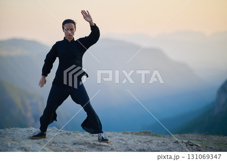 Woman practicing Tai Chi in traditional black outfit on mountain at sunrise, arms outstretched in meditation pose focusing on balance, energy, and mindfulness in nature. Woman practicing Tai Chi in traditional black outfit on mountain at sunrise, arms outstretched in meditation pose focusing on balance, energy, and mindfulness in nature. 131069347