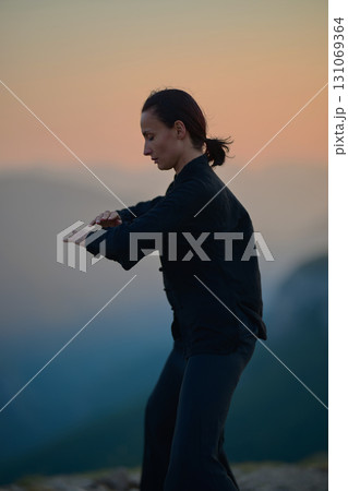 Woman practicing Tai Chi in traditional black outfit on mountain at sunrise, arms outstretched in meditation pose focusing on balance, energy, and mindfulness in nature. Woman practicing Tai Chi in traditional black outfit on mountain at sunrise, arms outstretched in meditation pose focusing on balance, energy, and mindfulness in nature. 131069364