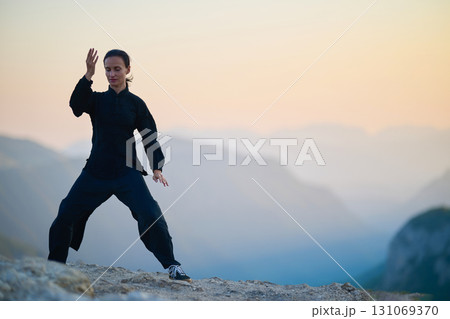 Woman practicing Tai Chi in traditional black outfit on mountain at sunrise, arms outstretched in meditation pose focusing on balance, energy, and mindfulness in nature. 131069370
