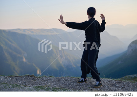 Woman practicing Tai Chi in traditional black outfit on mountain at sunrise, arms outstretched in meditation pose focusing on balance, energy, and mindfulness in nature. 131069425