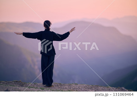 Woman practicing Tai Chi in traditional black outfit on mountain at sunrise, arms outstretched in meditation pose focusing on balance, energy, and mindfulness in nature. 131069464
