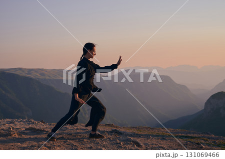 Woman practicing Tai Chi in traditional black outfit on mountain at sunrise, arms outstretched in meditation pose focusing on balance, energy, and mindfulness in nature. 131069466