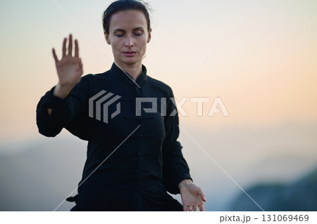 Woman practicing Tai Chi in traditional black outfit on mountain at sunrise, arms outstretched in meditation pose focusing on balance, energy, and mindfulness in nature. 131069469