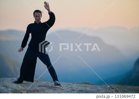 Woman practicing Tai Chi in traditional black outfit on mountain at sunrise, arms outstretched in meditation pose focusing on balance, energy, and mindfulness in nature. 131069472